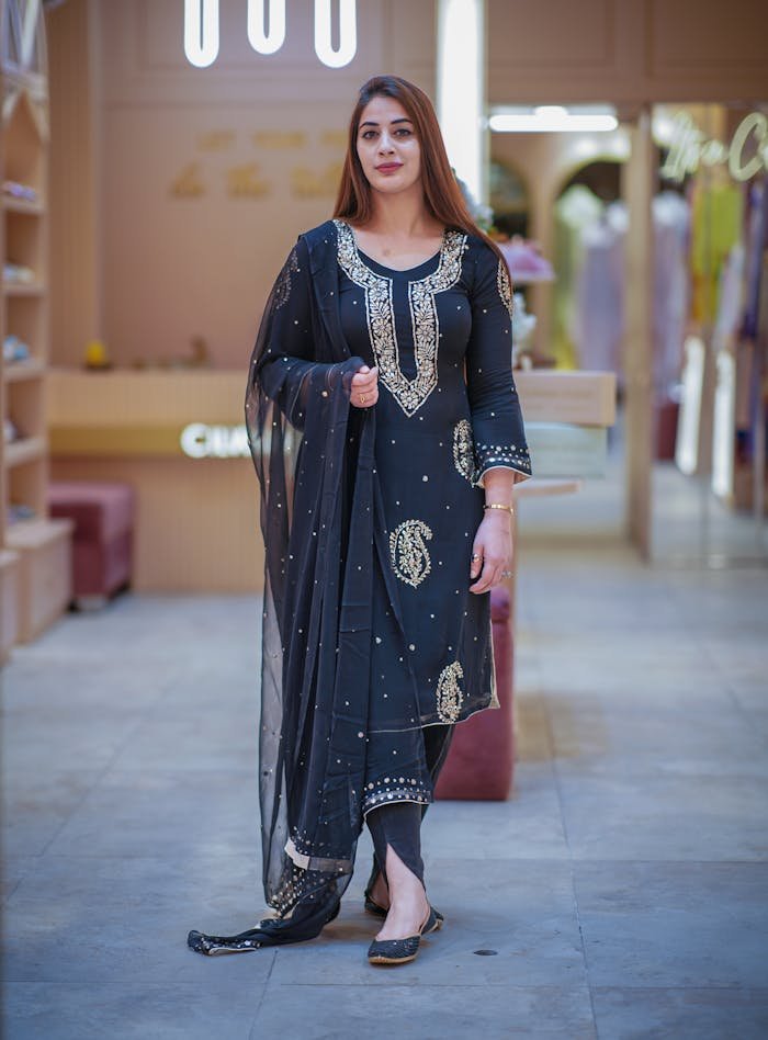 A stylish woman poses in a traditional black embroidered saree in a modern indoor setting.