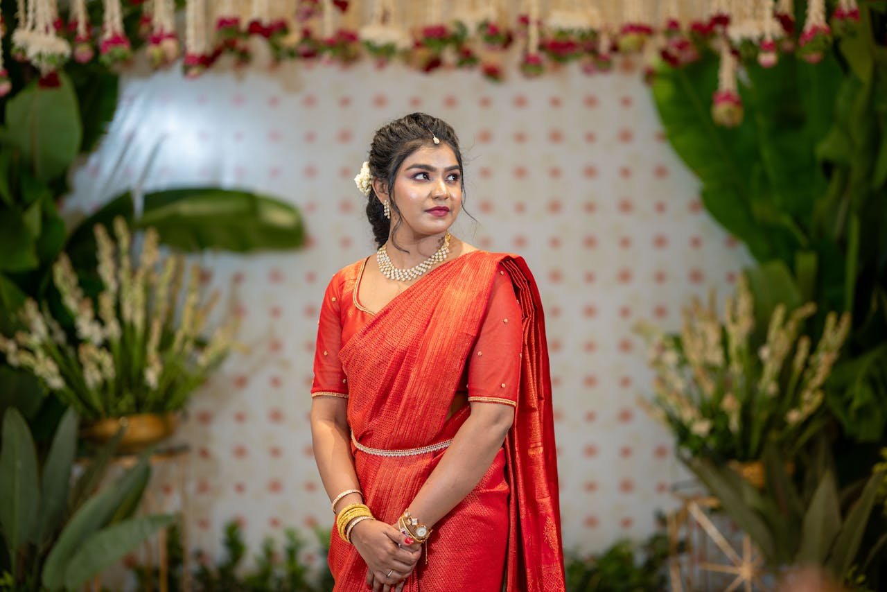 A woman in a vibrant red sari attends a formal indoor event with lush greenery.